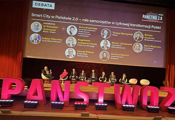 Na scenie siedzi osiem osób podczas debaty na temat roli samorządów w cyfrowej transformacji Polski, z dużym napisem PAŃSTWO 2.0 na pierwszym planie oraz wyświetlonymi imionami i stanowiskami uczestników debaty na tle ekranu.