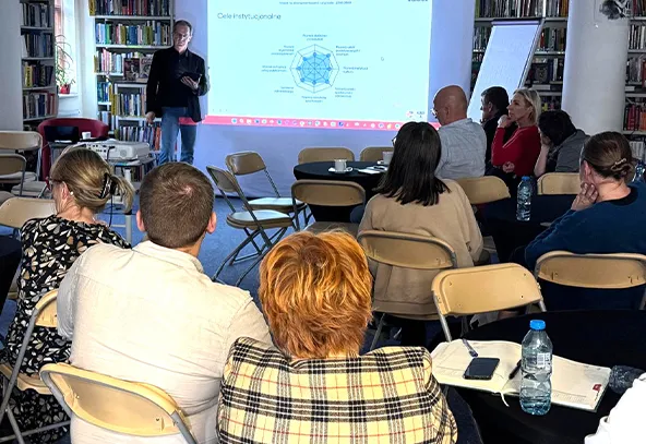 Grupa osób siedzi w kręgu na krzesłach podczas prezentacji w bibliotece. Mężczyzna stoi przy ekranie z wyświetlonym wykresem. W tle widać regały z książkami.