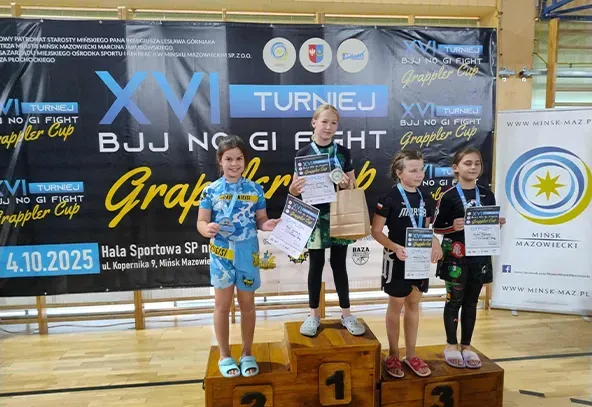 Cztery dziewczynki stoją na drewnianej podium w hali sportowej. Najwyższe miejsce zajmuje dziewczynka z medalem i dyplomem. Pozostałe trzy dziewczynki również trzymają dyplomy i medale. W tle znajduje się baner z informacjami o turnieju Grappler Cup. Podłoga drewniana, wnętrze jasne i dobrze oświetlone.