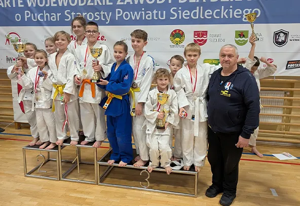 Grupa dzieci w strojach do judo stoi na podium w hali sportowej, kilka osób trzyma puchary i medale, obok nich stoi dorosły mężczyzna w ciemnej bluzie, w tle widać baner z tekstem i logotypami.