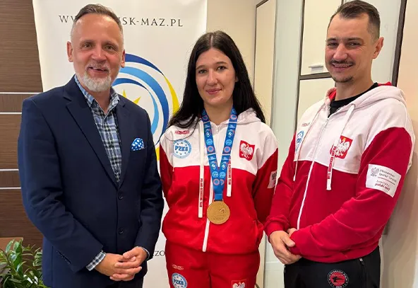 Trzy osoby stoją wewnątrz pomieszczenia przy jasnej ścianie z banerem w tle. Kobieta pośrodku ma na szyi złoty medal i ubrana jest w biało-czerwony strój sportowy z emblematem Polski. Mężczyźni po bokach uśmiechają się, jeden ma niebieską marynarkę, drugi biało-czerwony strój sportowy.