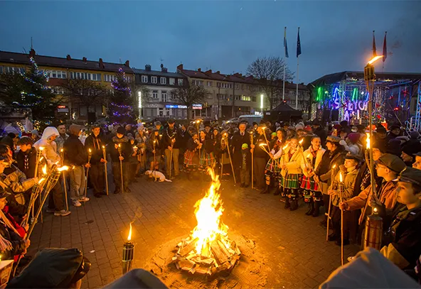 Ludzie stoją wokół ogniska na placu w czasie wieczoru. Część osób trzyma pochodnie. W tle iluminacje świąteczne i budynki mieszkalne. Niebo jest ciemne, zbliża się noc.