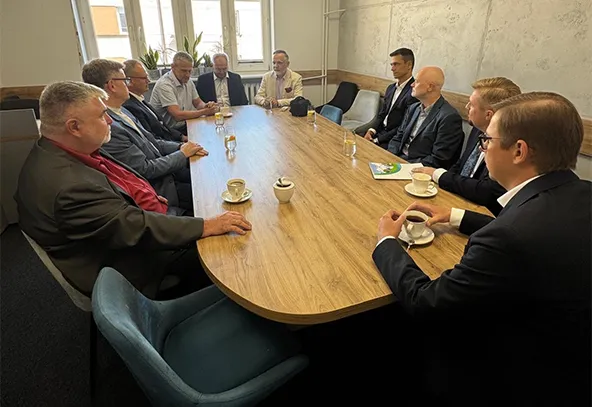 Dziewięciu mężczyzn w garniturach siedzi przy długim drewnianym stole w jasnym pomieszczeniu podczas spotkania, na stole są filiżanki i szklanki z napojami.