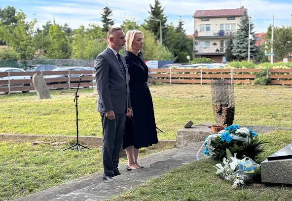 Mężczyzna i kobieta stoją na zewnątrz przy pomniku z wieńcami i kwiatami w czasie dnia.