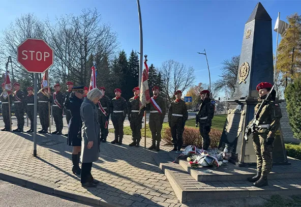 Grupa żołnierzy w mundurach i czerwonych beretach stoi na chodniku przy pomniku z kwiatami. Dwie osoby w cywilnych strojach stoją przodem do pomnika. W tle widać drzewa oraz znak drogowy STOP. Jest dzień, niebo jest bezchmurne.