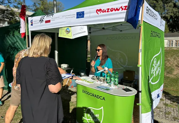 Stoisko z zielonymi ściankami i parasolem na zewnątrz, przy którym stoi kobieta z mikrofonem i rozmawia z dwoma osobami. Na stoisku leżą butelki i ulotki. W tle drzewa i budynek.