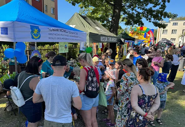 Duża grupa ludzi stoi na zewnątrz przy namiotach podczas słonecznego dnia, obok są budynki mieszkalne i kolorowa dmuchana zjeżdżalnia.