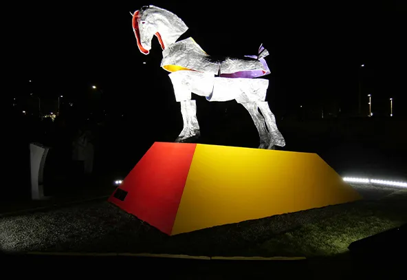 Posąg konia wykonanego z jasnego materiału umieszczony na prostokątnej podstawie pomalowanej na czerwono i żółto, ustawiony na zewnątrz nocą na trawniku.