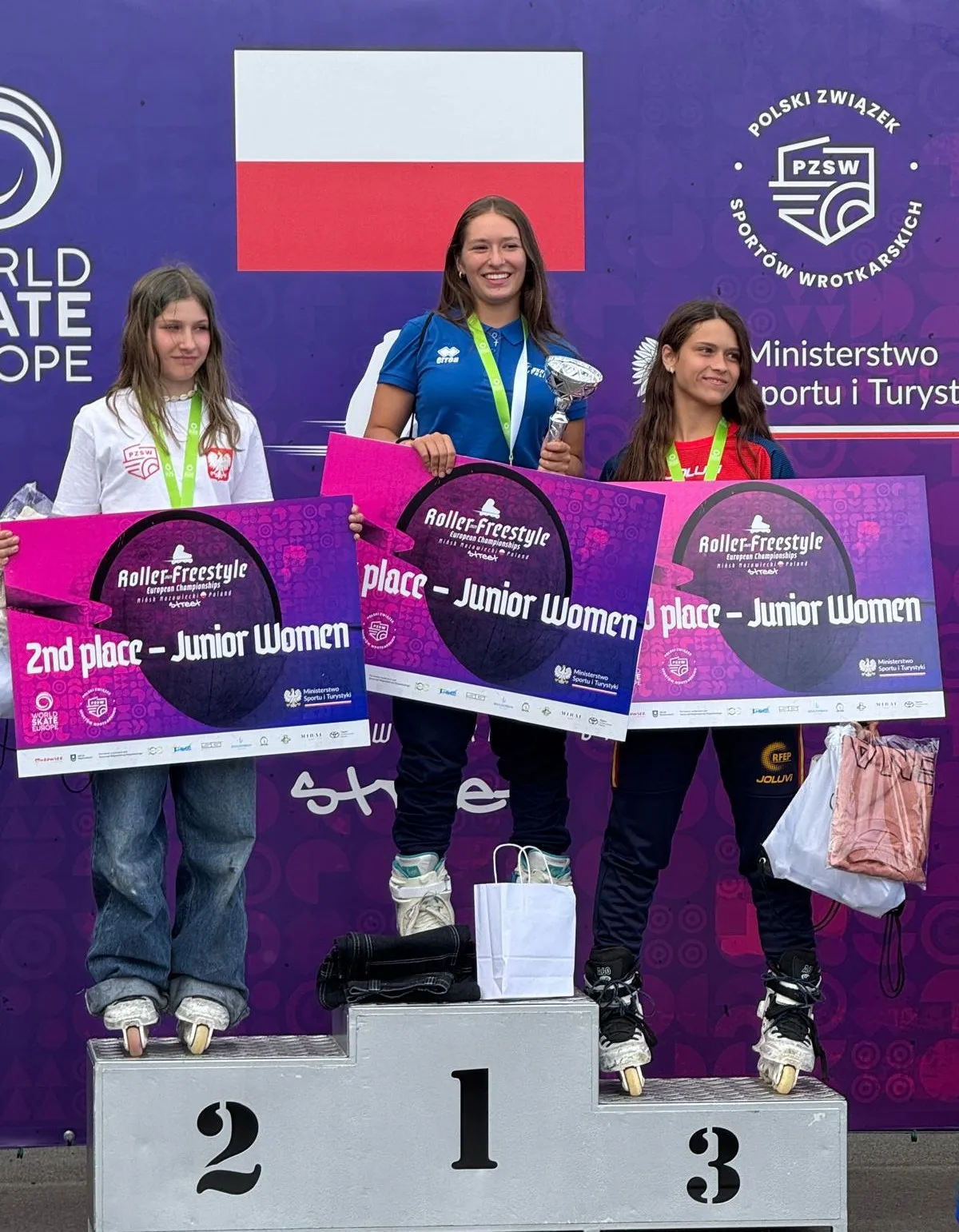 Trzy zawodniczki na podium z medalami i dyplomami za miejsca w konkurencji Junior Women na Roller-Freestyle European Championships przed flagą Polski i banerem PZSW oraz Ministerstwa Sportu i Turystyki.