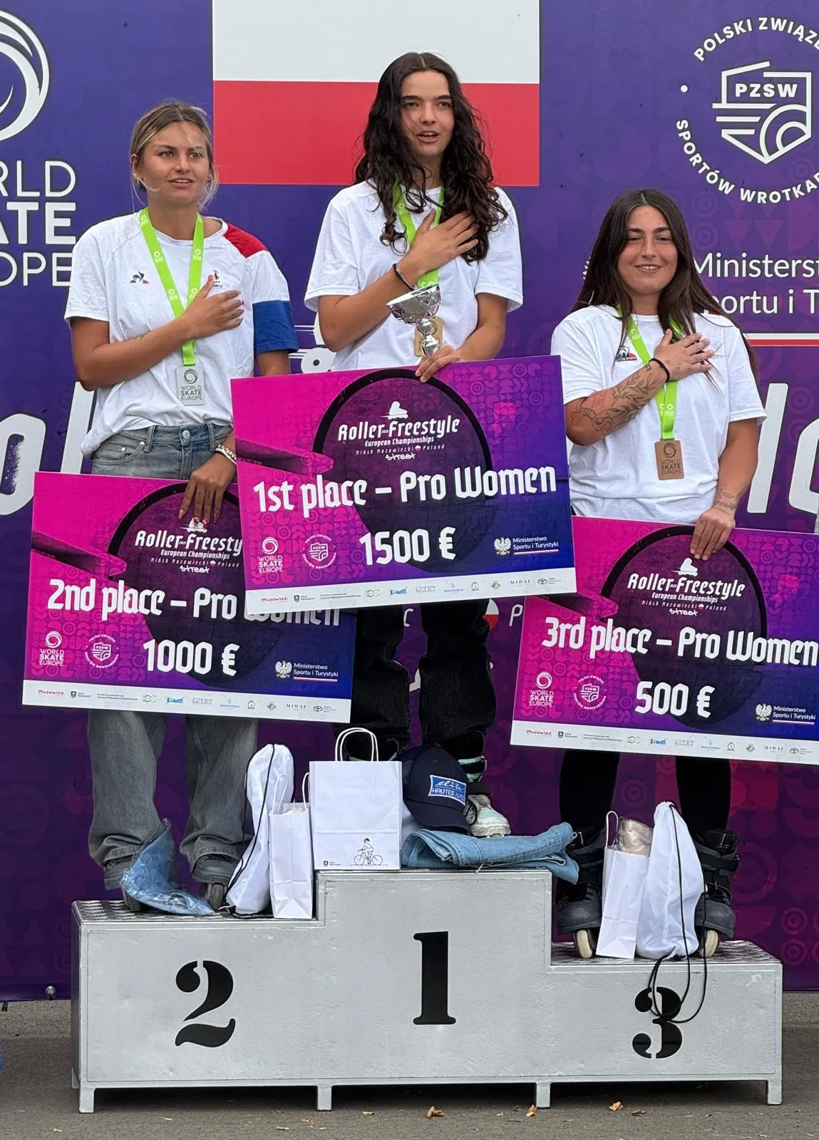 Trzy kobiety na podium trzymające tablice z nagrodami za 1., 2. i 3. miejsce w zawodach Roller-Freestyle European Championships, pierwsza miejsce na środku, wszystkie mają na szyjach medale i trzymają rękę na sercu.