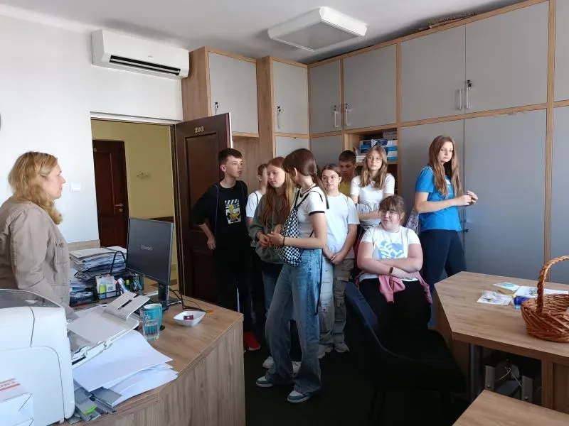 Grupa młodzieży stoi w pokoju z biurkiem, komputerem i szafkami, a kobieta stoi naprzeciwko nich.