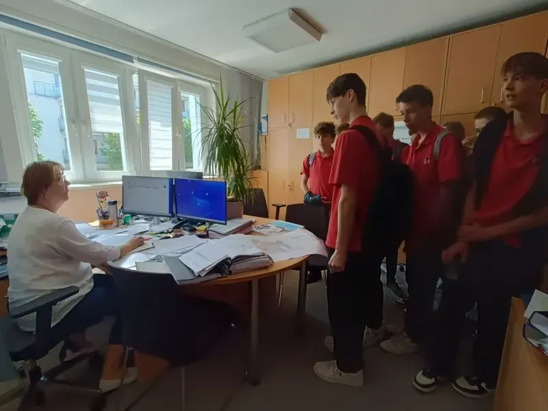 Nauczycielka siedzi przy biurku z dwoma monitorami, a grupa uczniów w czerwonych koszulkach stoi naprzeciwko niej.