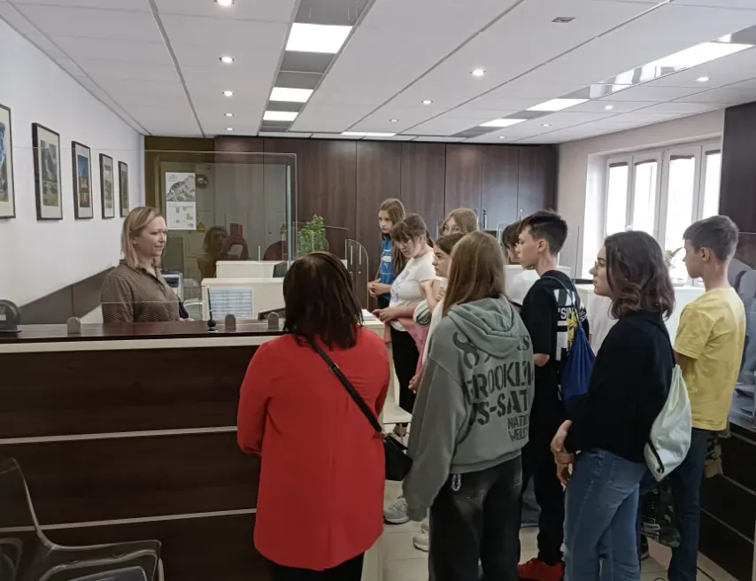 Grupa młodych osób stoi przy ladzie w biurze, rozmawiając z kobietą za osłoną z pleksi.