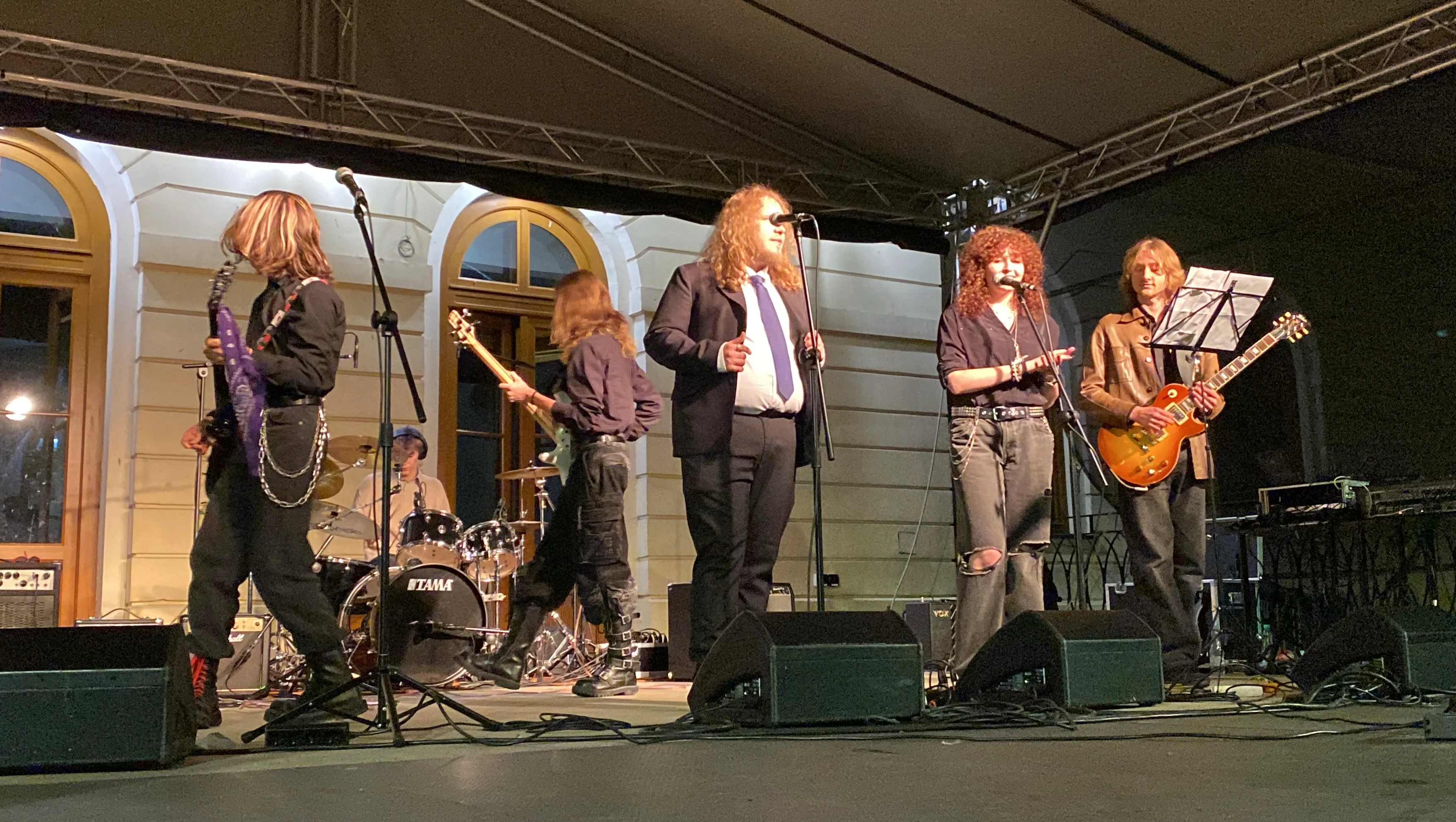 Sześć osób na scenie podczas koncertu na zewnątrz w nocy, trzech mężczyzn gra na gitarach, jedna osoba siedzi przy perkusji, dwie osoby stoją przy mikrofonach, budynek oświetlony w tle.