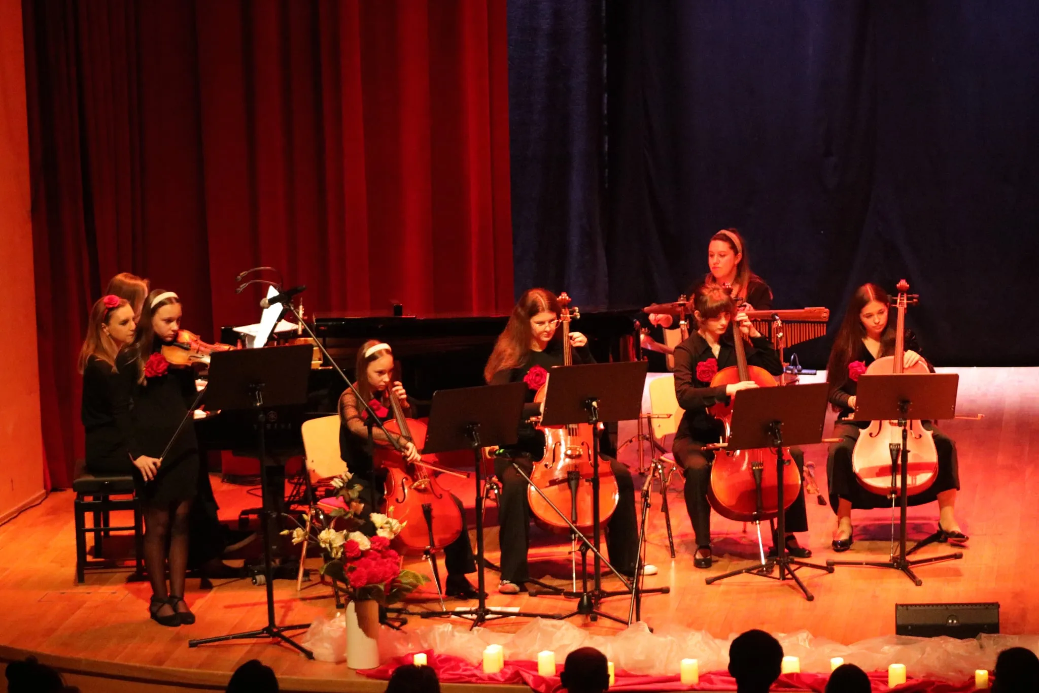 Siedem młodych kobiet na scenie gra na instrumentach smyczkowych i perkusji, ubranych na czarno z czerwonymi akcentami. Za nimi czerwone i granatowe zasłony. Na scenie stoją kwiaty i zapalone świece. Publiczność siedzi na widowni.
