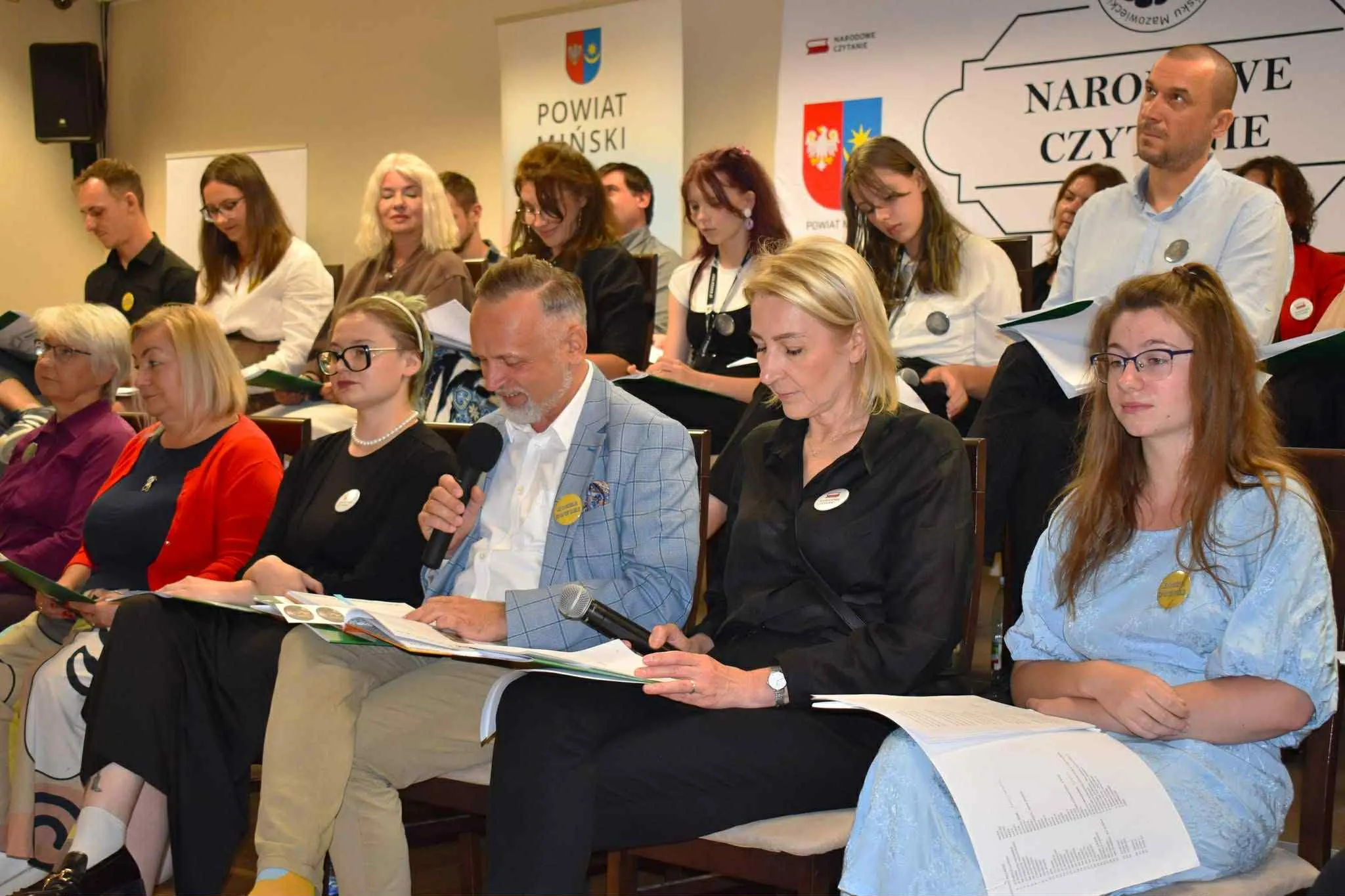 Grupa dorosłych i młodych osób siedzi na krzesłach w sali podczas wydarzenia związanego z czytaniem, niektórzy trzymają mikrofony i czytają z książek lub kartek.