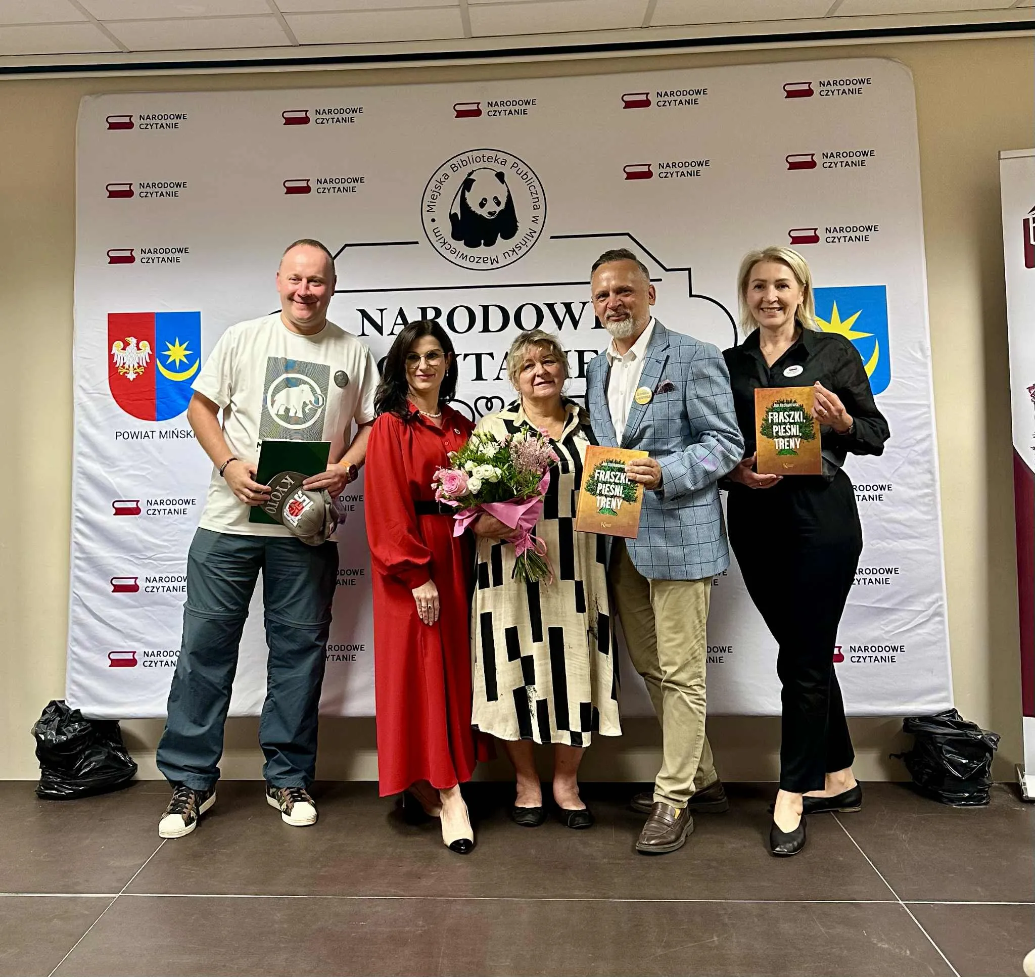 Pięć osób stoi na tle baneru z napisem Narodowe Czytanie i logo Miejskiej Biblioteki Publicznej w Mińsku Mazowieckim, dwie z nich trzymają książki, a jedna bukiet kwiatów.