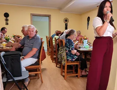 Kobieta trzyma mikrofon, a grupa starszych osób siedzi przy stołach w jadalni.