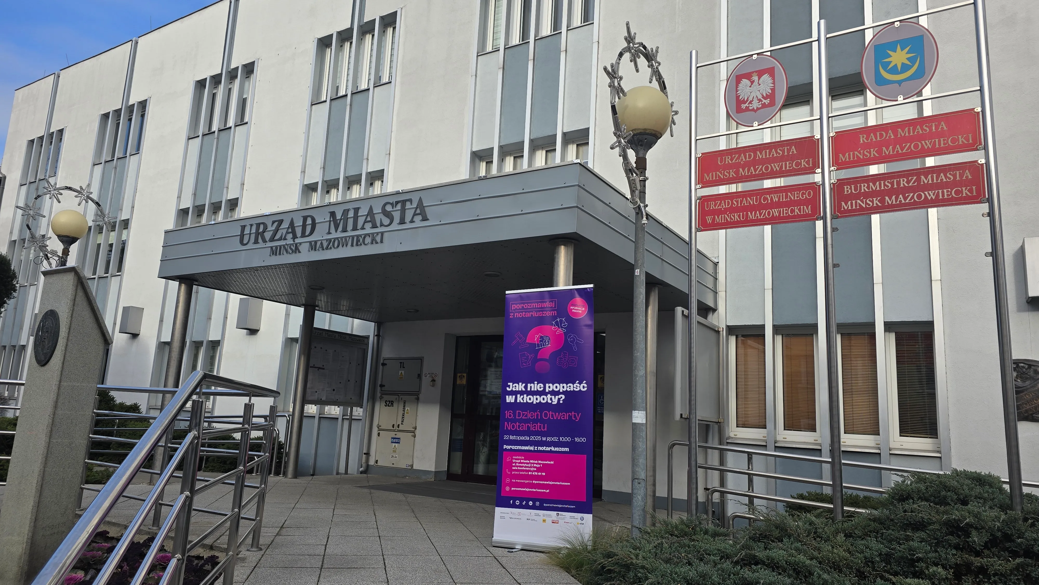 Wejście do budynku Urzędu Miasta Mińsk Mazowiecki w dzień, z oszklonymi drzwiami i dwoma lampami ulicznymi po bokach. Po prawej stronie tablice z herbami i nazwami instytucji na czerwonym tle. Przed wejściem stoi stojak z plakatem informacyjnym na fioletowym tle. Na pierwszym planie metalowa barierka i niska roślinność.