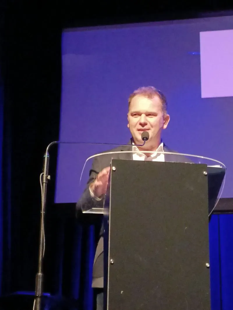 Mężczyzna w garniturze stoi za mównicą z mikrofonem i przemawia na tle niebieskiego ekranu.