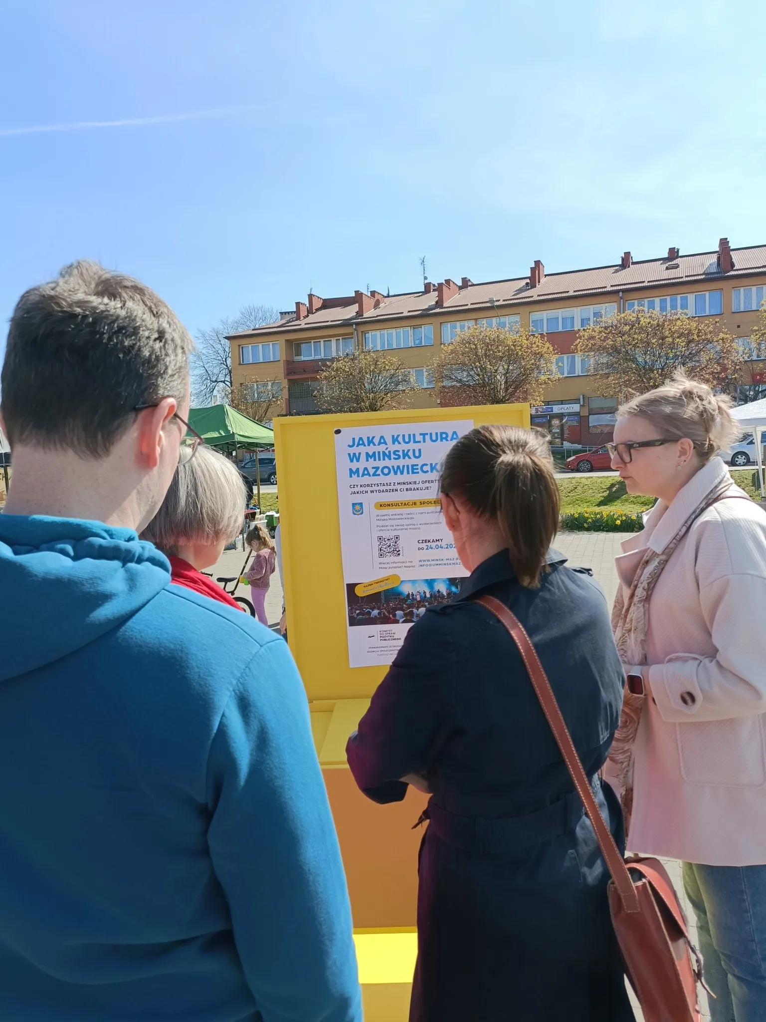 Cztery osoby stoją na zewnątrz przy żółtym stoisku z plakatem dotyczącym kultury w Mińsku Mazowieckim, w tle budynki mieszkalne i zielony teren.