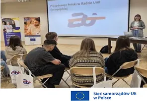Kobieta przemawia do grupy siedzących osób na sali, na ścianie wyświetlany jest slajd z napisem dotyczącym Polski w Radzie Unii Europejskiej.