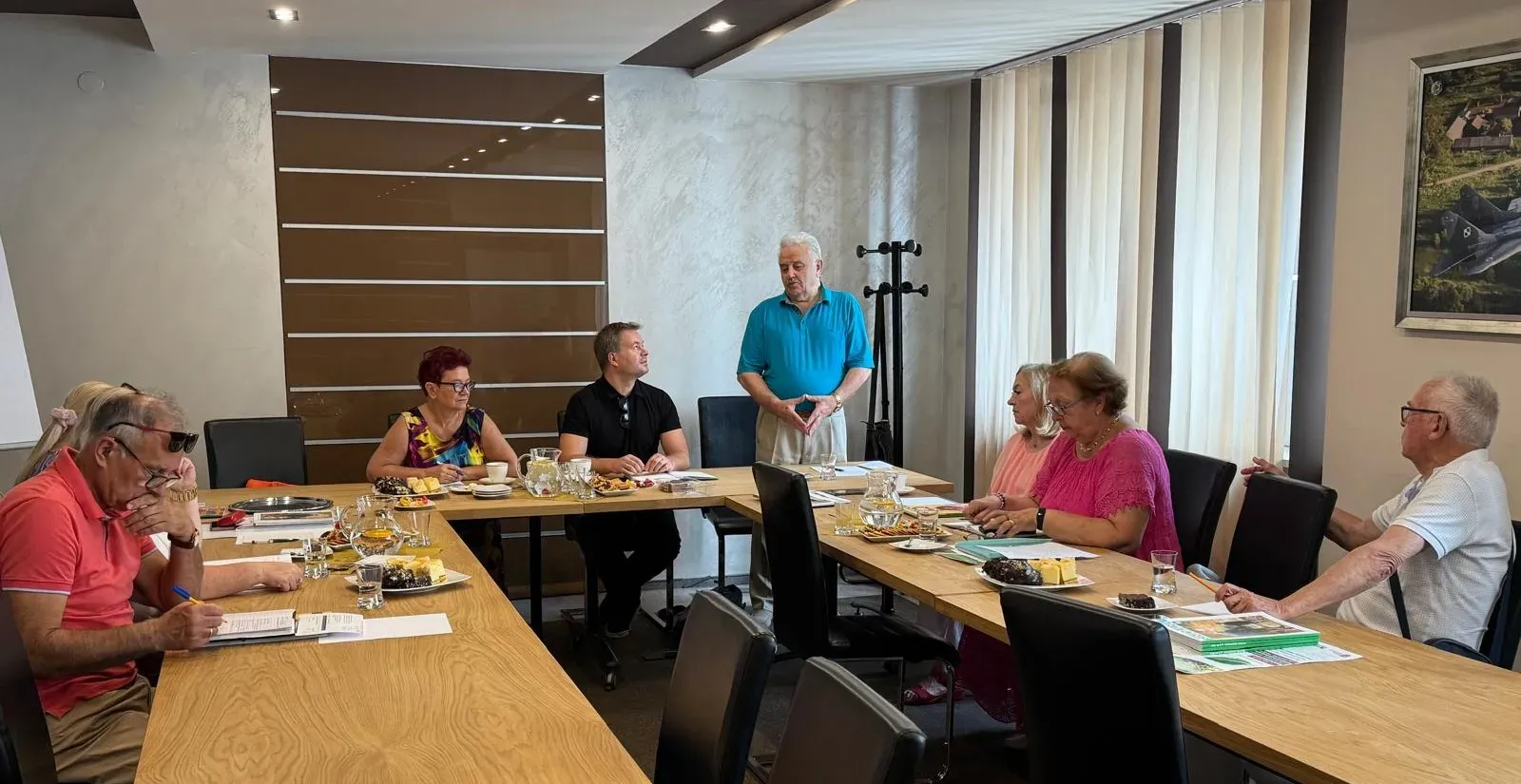 Osiem osób podczas spotkania w sali konferencyjnej, mężczyzna w niebieskiej koszulce stoi przy stole, reszta osób siedzi i słucha, na stole dokumenty, talerze z ciastem i dzbanki z napojem.