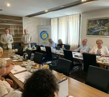 Grupa osób siedzących przy długim stole konferencyjnym z dokumentami, filiżankami i butelkami wody, w sali z dużymi oknami i roletami.