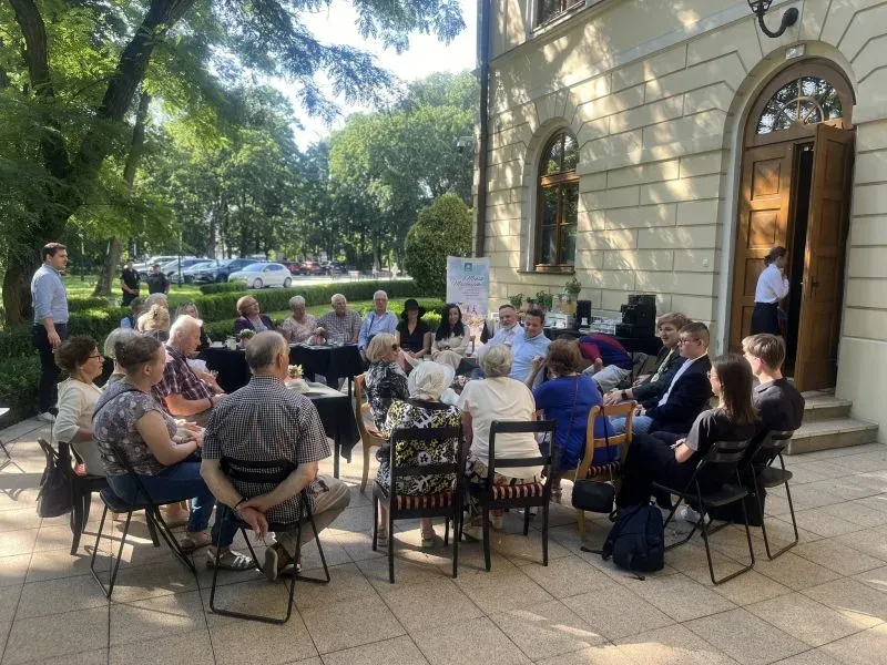 Grupa osób siedzi na krzesłach na dziedzińcu przed budynkiem i uczestniczy w spotkaniu.