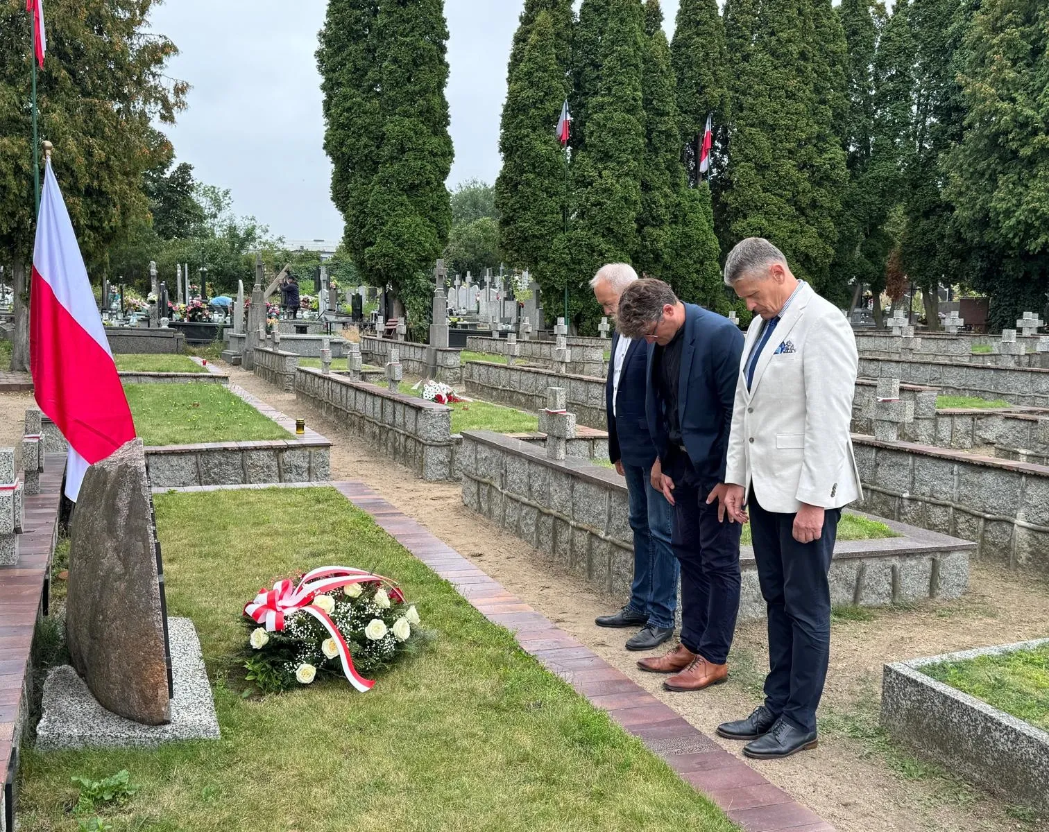 Trzech mężczyzn stoi na cmentarzu obok grobu z wieńcem i flagą Polski, pochylając się w zadumie.