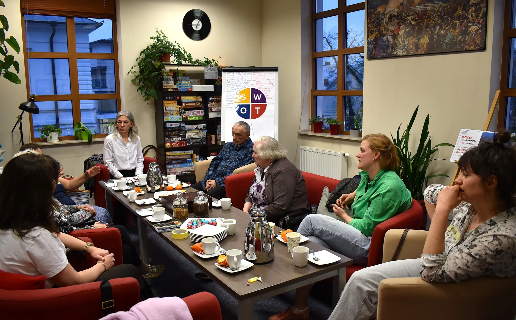 Grupa ośmiu osób siedzi przy stole w pokoju z dużymi oknami, na stole filiżanki, dzbanki, przekąski, a na ścianie plansza z analizą SWOT.