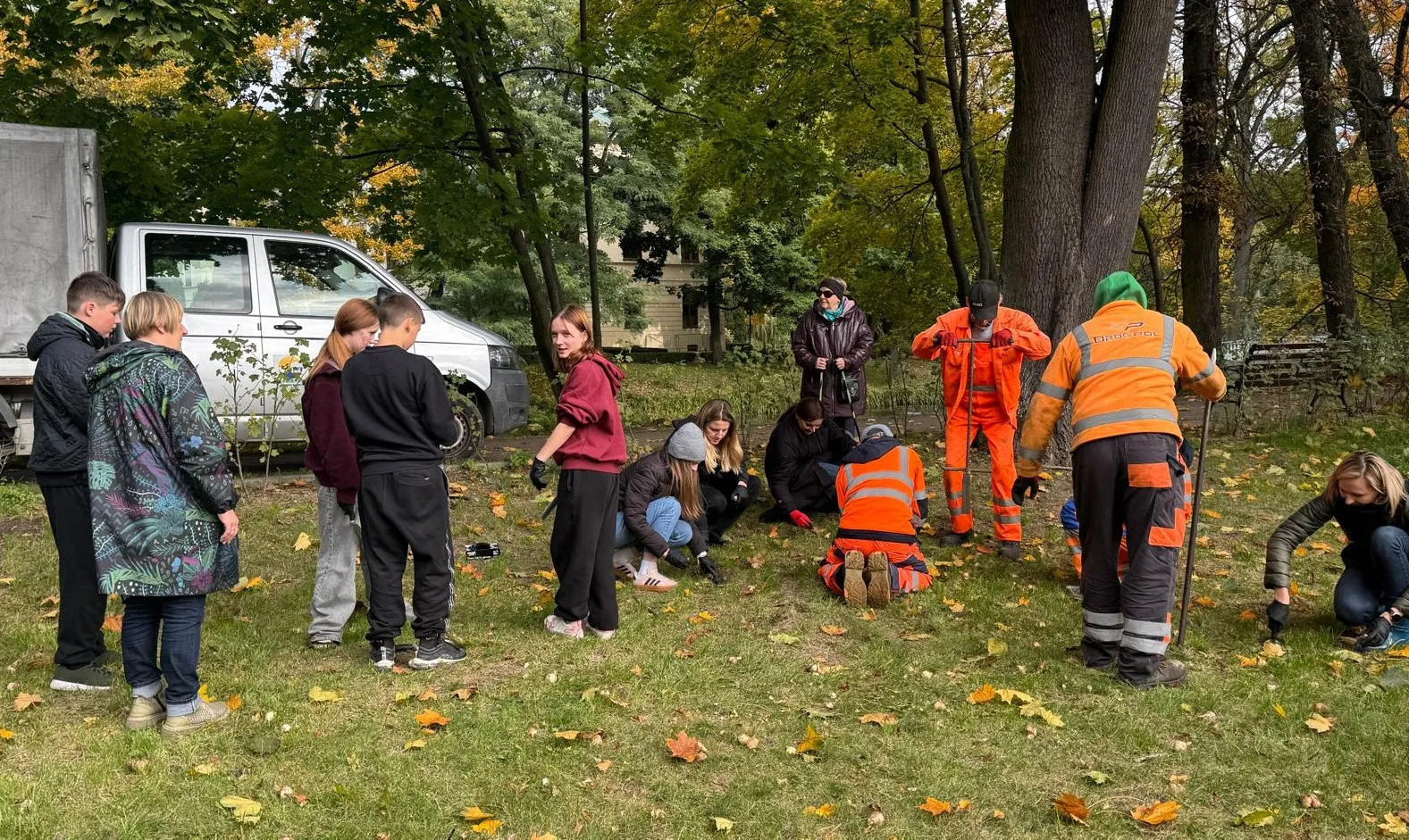 Grupa ludzi na trawie w parku jesienią. Kilka osób klęczy i pracuje przy ziemi, dwie osoby ubrane w pomarańczowe ubrania robocze, inni stoją lub kucają. W tle białe auto dostawcze oraz drzewa z zielonymi i żółtymi liśćmi.