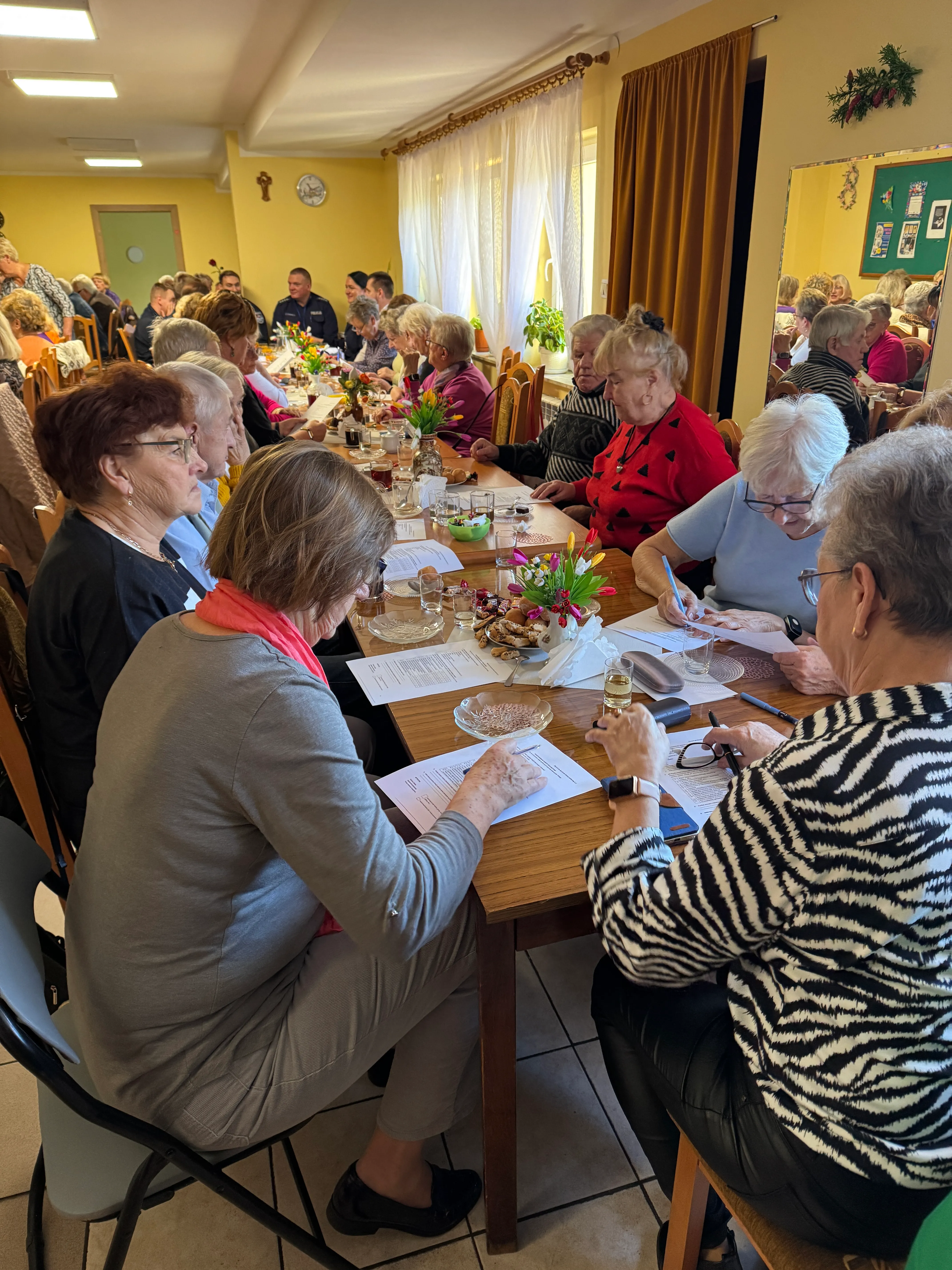 Grupa około dwudziestu osób, głównie starszych kobiet, siedzi przy długim stole w jasnym pomieszczeniu. Na stole znajdują się dokumenty, kwiaty w wazonach oraz napoje. Za oknem widoczne są białe firany i żółte ściany. W tle widoczna jest tablica informacyjna, lustro i kilka dekoracji ściennych.
