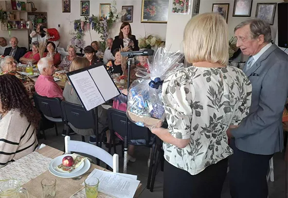 Grupa dorosłych osób siedzi przy stołach w pomieszczeniu, kobieta trzyma kosz prezentowy stoi obok starszego mężczyzny przy mikrofonie.