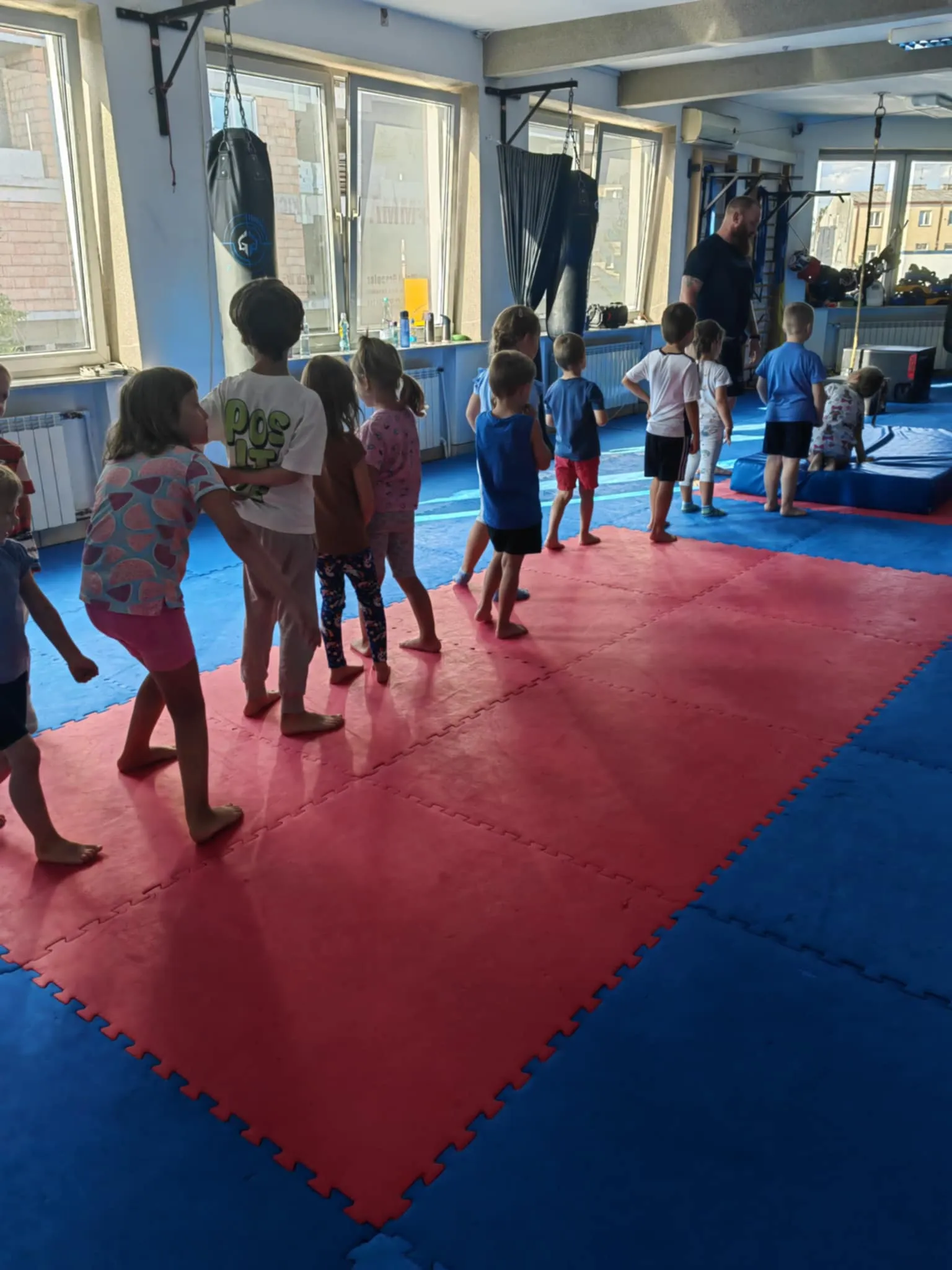 Grupa dzieci ustawia się w kolejce na macie w sali gimnastycznej z instruktorem.