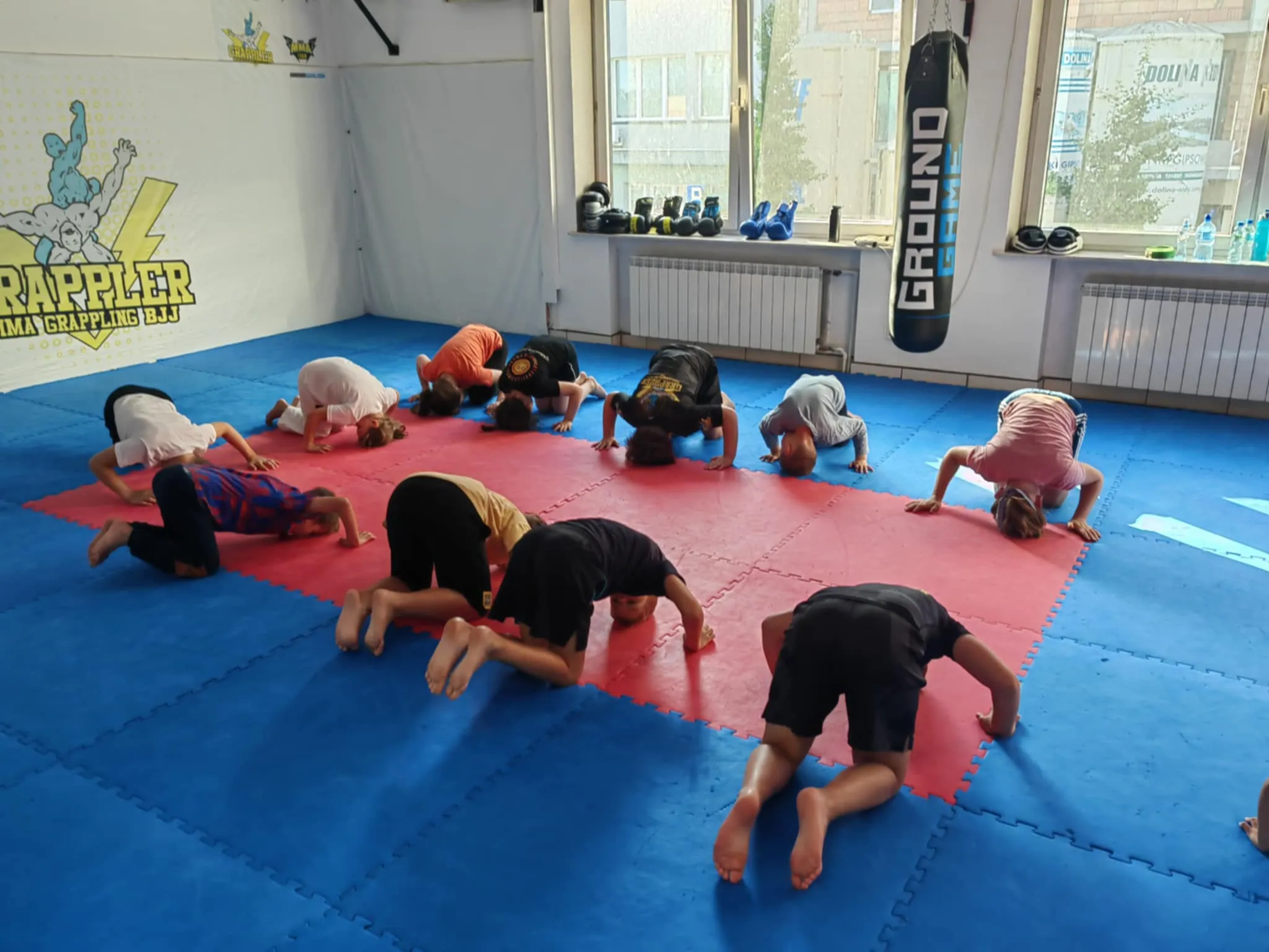 Dziesięcioro dzieci ćwiczy na matach w sali do grapplingu, wykonując ćwiczenie na czworakach.