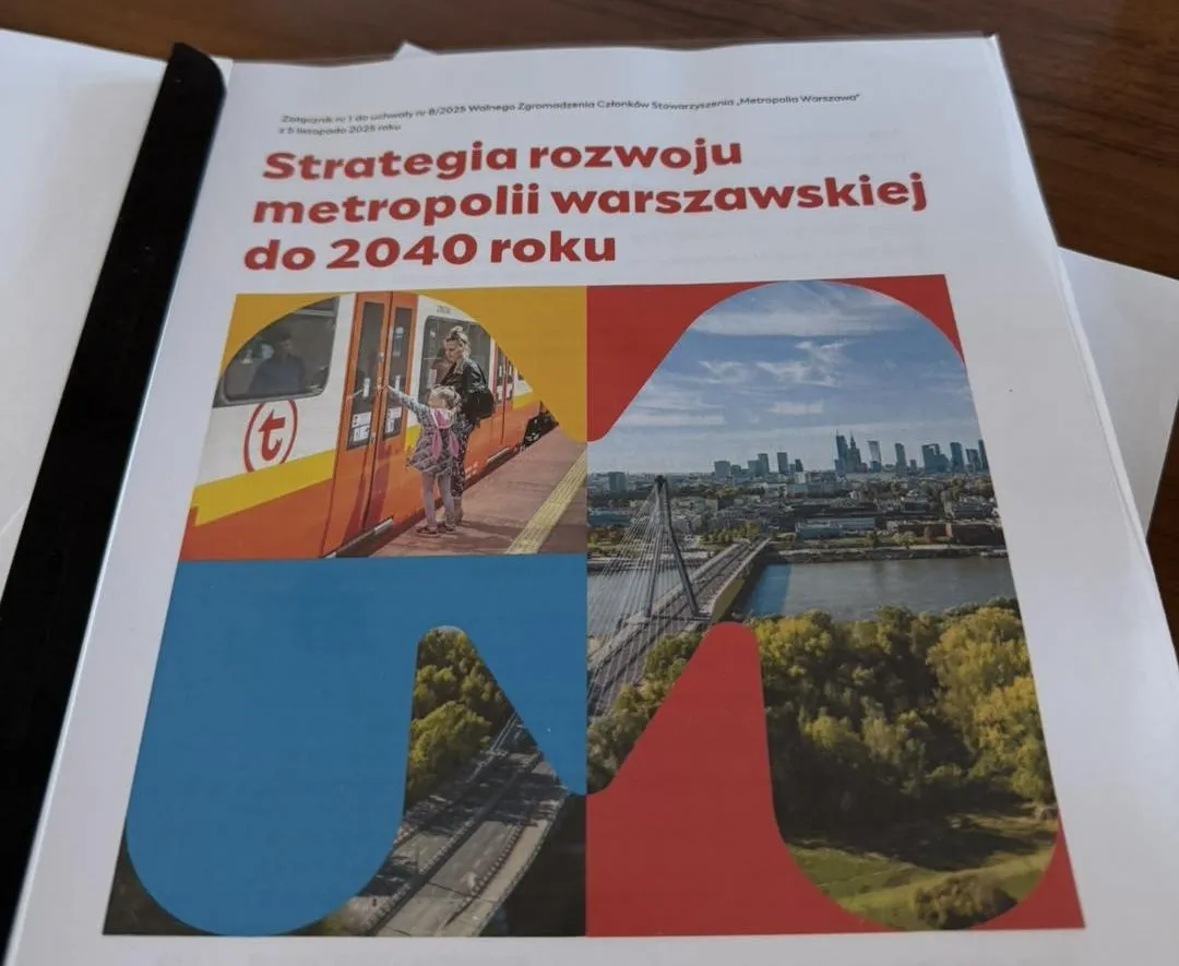 Dokument leżący na drewnianym stole z dużym napisem „Strategia rozwoju metropolii warszawskiej do 2040 roku” oraz grafiką w kształcie litery „M” zawierającą zdjęcia: kobietę z dzieckiem wsiadającą do pociągu, panoramę Warszawy z rzeką i mostem oraz zdjęcie torów kolejowych z zielenią.