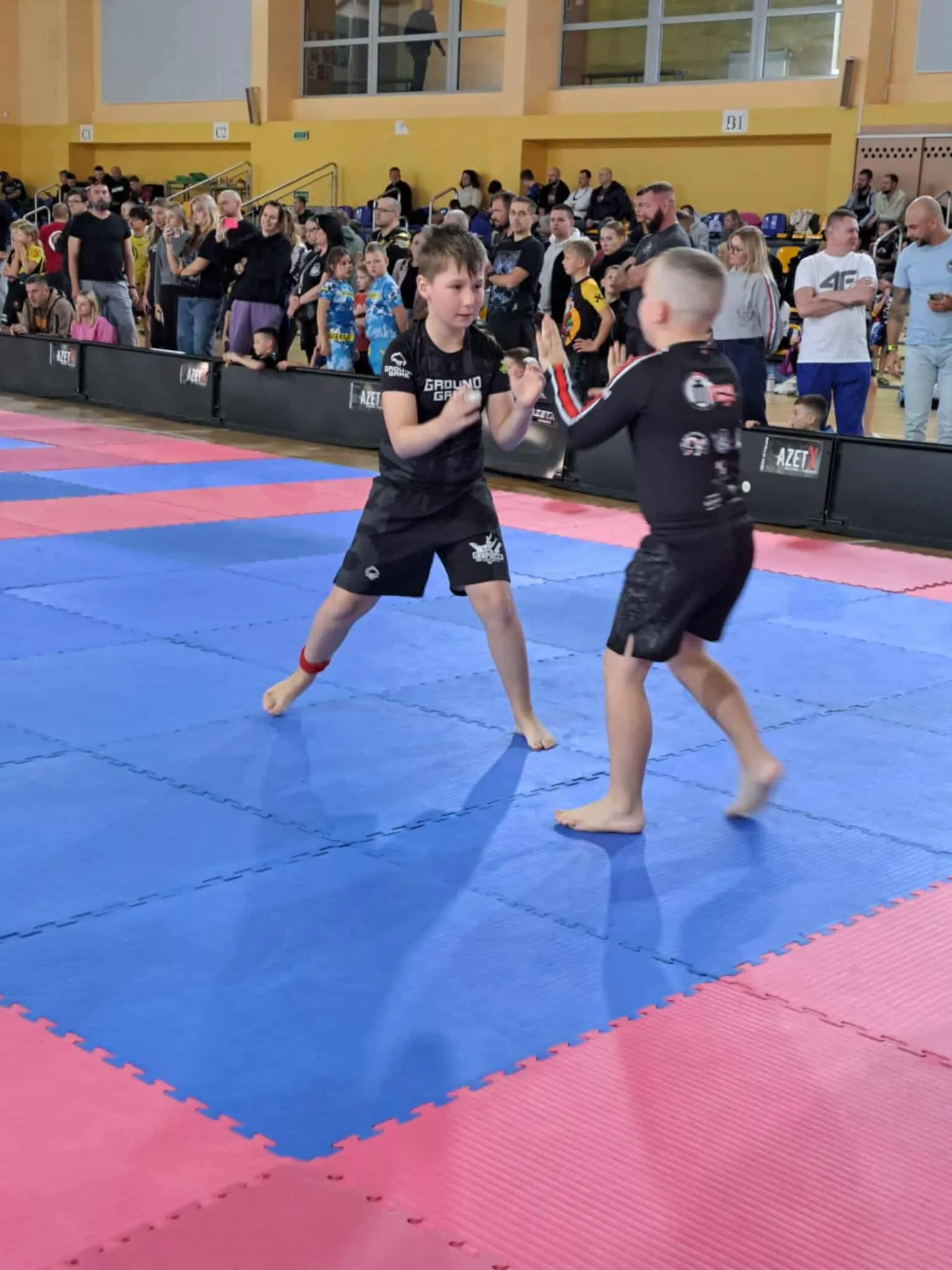 Dwóch chłopców trenuje sporty walki na niebiesko-różowej macie w hali sportowej. W tle stoi grupa osób obserwujących trening.