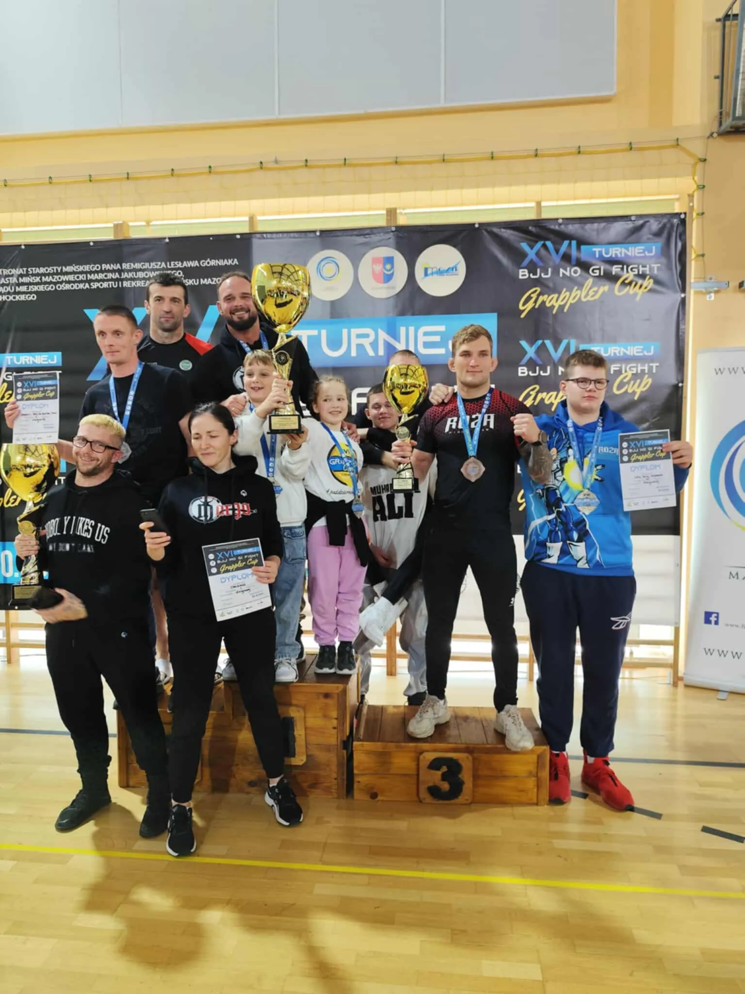Grupa dziesięciu osób, w tym dwóch dzieci, stoi na podium i na parkiecie hali sportowej. Niektórzy trzymają duże złote puchary i dyplomy, wszyscy mają medale na szyjach. W tle wisi duży czarny baner z napisem „XVI TURNIEJ BJJ NO GI FIGHT Grappler Cup”.