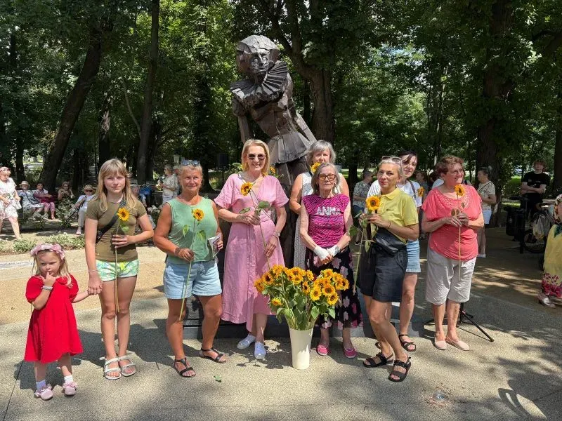 Grupa kobiet i dziewczynek stoi przed dużą rzeźbą kobiecej postaci. Każda trzyma słonecznik. Na ziemi stoi bukiet słoneczników.