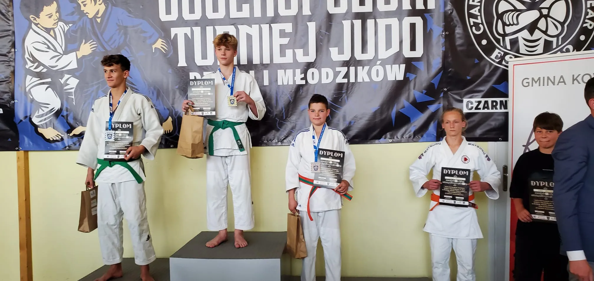 Pięciu zawodników na podium podczas turnieju judo, każdy trzyma dyplom i torbę, w tle baner z napisem o turnieju.