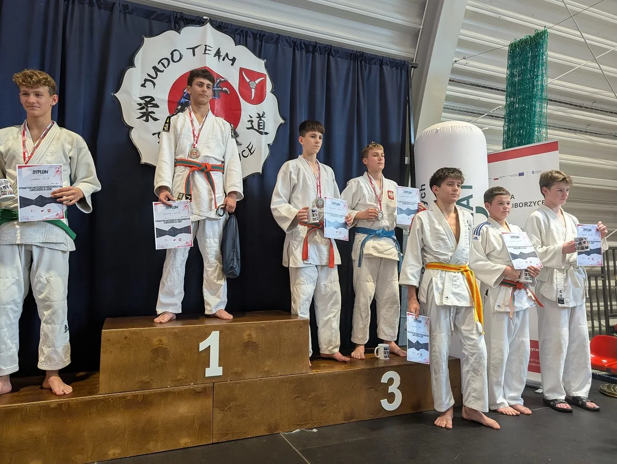 Siedmiu nastoletnich zawodników w judogach stoi na podium i obok niego w hali sportowej. Trzech z nich stoi na podium oznaczonym numerami 1 i 3, pozostali obok. Wszyscy trzymają dyplomy i mają na szyjach medale. Za nimi wisi granatowa kotara z logo klubu judo.