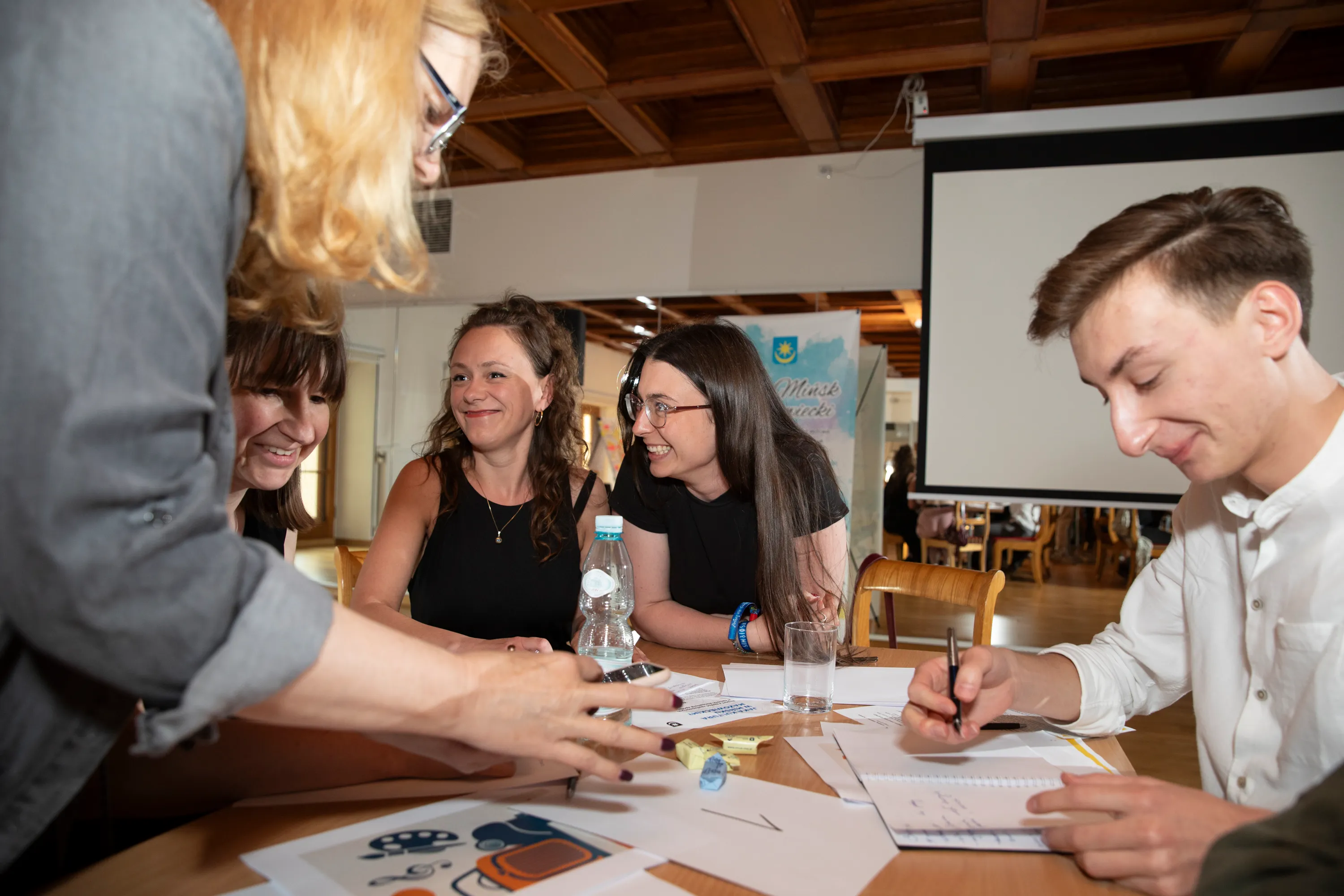 Pięć osób siedzi przy stole z dokumentami i pisakiem, rozmawiając i uśmiechając się podczas spotkania w jasnym wnętrzu z drewnianym sufitem.