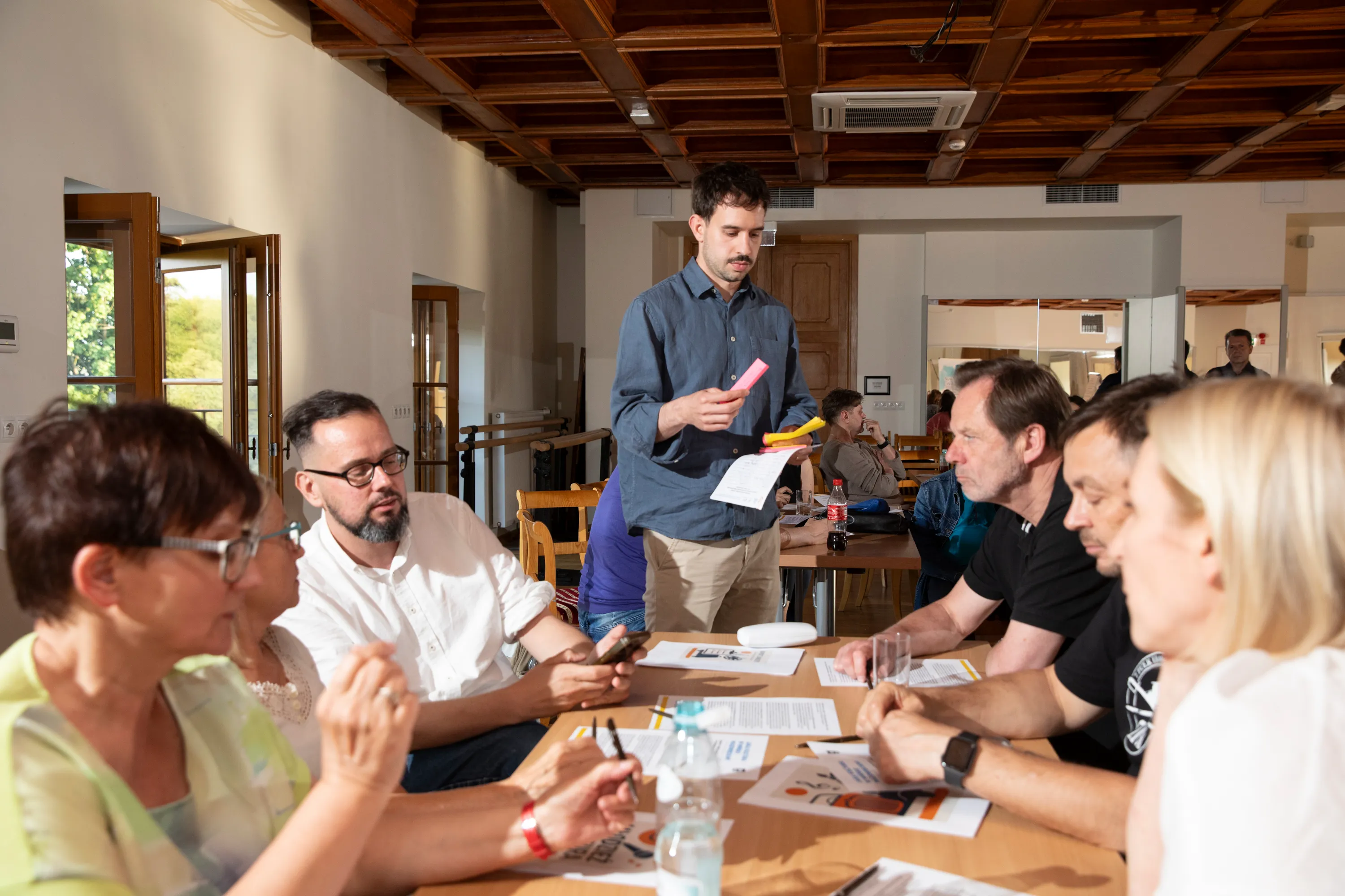 Grupa siedmiu dorosłych osób siedzi przy stole w jasnym pokoju podczas spotkania, a mężczyzna stojący w środku trzyma kolorowe karteczki i kartkę papieru.