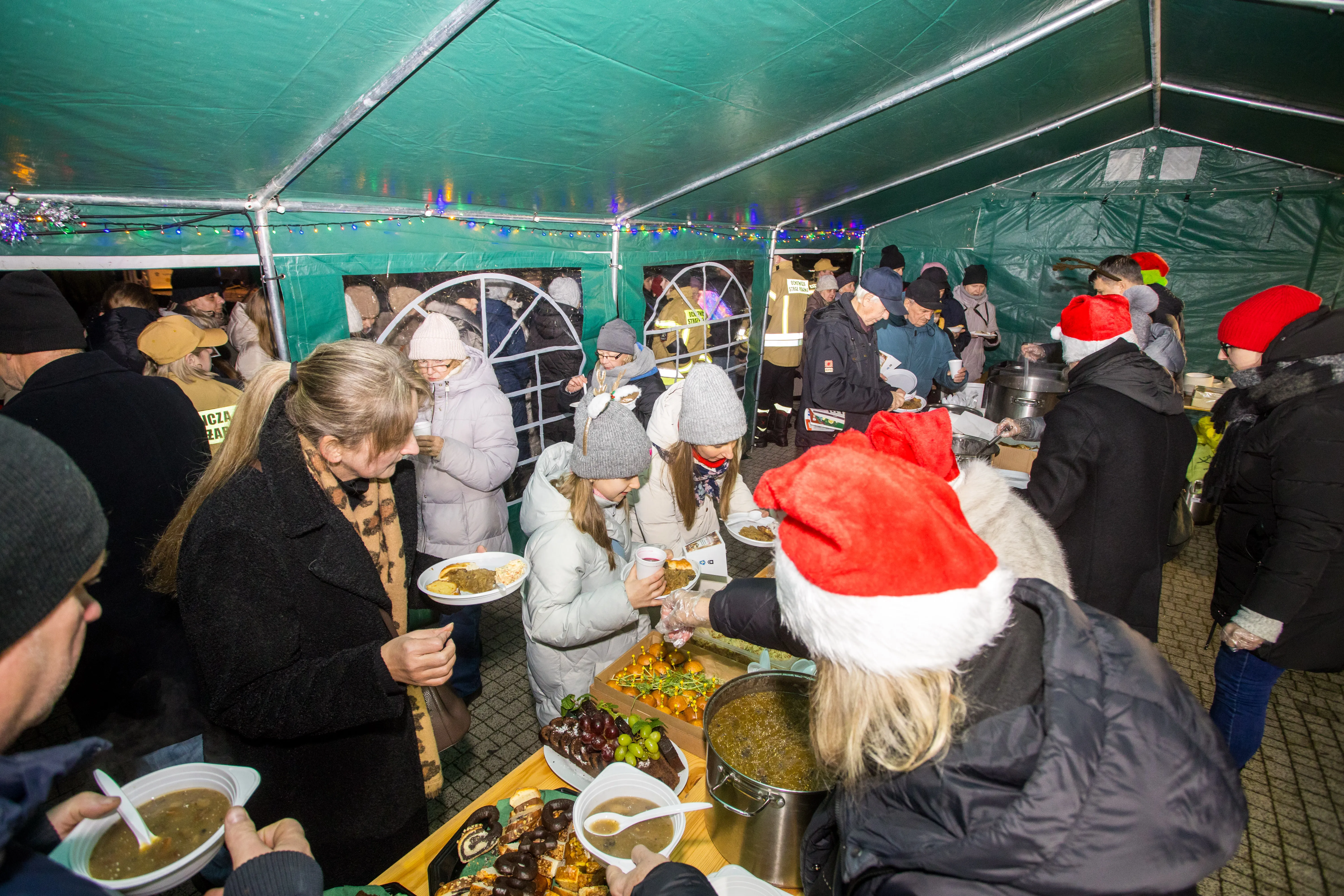 Grupa ludzi w ciepłych ubraniach stoi pod zielonym namiotem. Kilka osób w czerwonych czapkach Świętego Mikołaja serwuje jedzenie z dużych garnków. Na stole leżą owoce i ciasta. W tle świecą kolorowe lampki. Wydarzenie odbywa się wieczorem na zewnątrz.