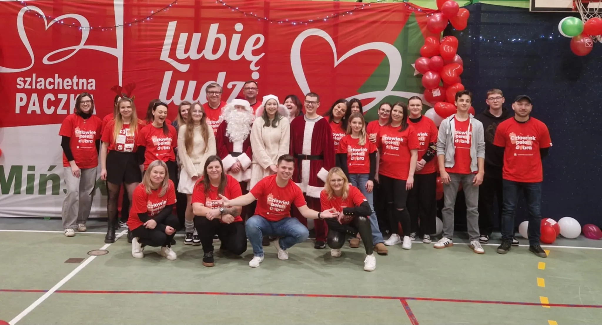 Grupa dwudziestu osób, z których większość ma czerwone koszulki z napisem, stoi i kuca na hali sportowej przed dużym czerwono-białym banerem z napisem „Szlachetna Paczka” i „Lubię ludzi”. W centrum dwie osoby przebrane za Świętego Mikołaja i kobietę w białym płaszczu oraz kilka osób w jasnych ubraniach. Po prawej stronie balony w kolorze czerwonym, zielonym i białym.