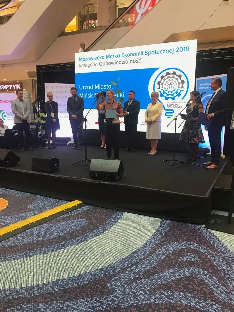 Grupa osób stojących na scenie podczas wręczenia nagrody Mazowiecka Marka Ekonomii Społecznej 2019, z kobietą trzymającą mikrofon i dyplom na pierwszym planie.