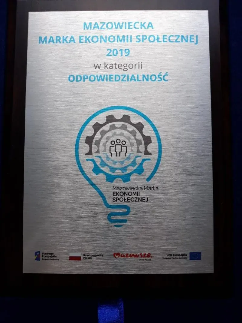 Tabliczka z napisem „Mazowiecka Marka Ekonomii Społecznej 2019 w kategorii Odpowiedzialność” oraz grafiką żarówki zębatki i sylwetkami ludzi.