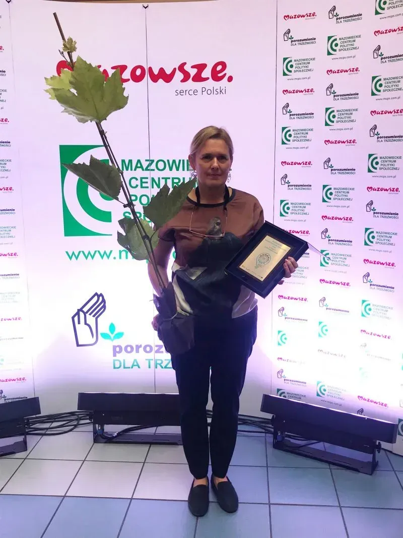 Kobieta stoi na tle bannera z logo Mazowsze i Mazowieckiego Centrum Polityki Społecznej, trzymając w jednej ręce doniczę z rośliną, a w drugiej oprawioną nagrodę.