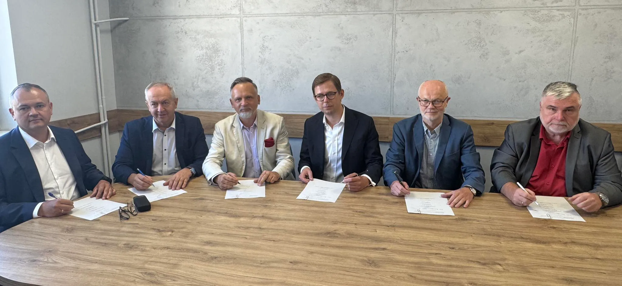 Sześciu mężczyzn siedzi przy drewnianym stole przy ścianie, podpisując dokumenty w jasnym, biurowym pomieszczeniu.